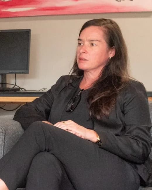 Portrait of Elaine French in a dark jacket, seated on a grey sofa