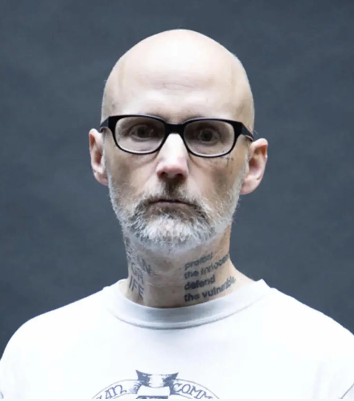 Portrait of Moby in black glasses and a white T-shirt against a dark grey backdrop