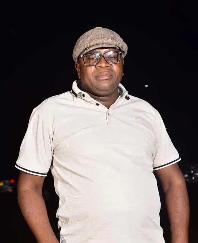 Portrait of Elmon Mudenda in a flat cap, glasses and white polo shirt at night