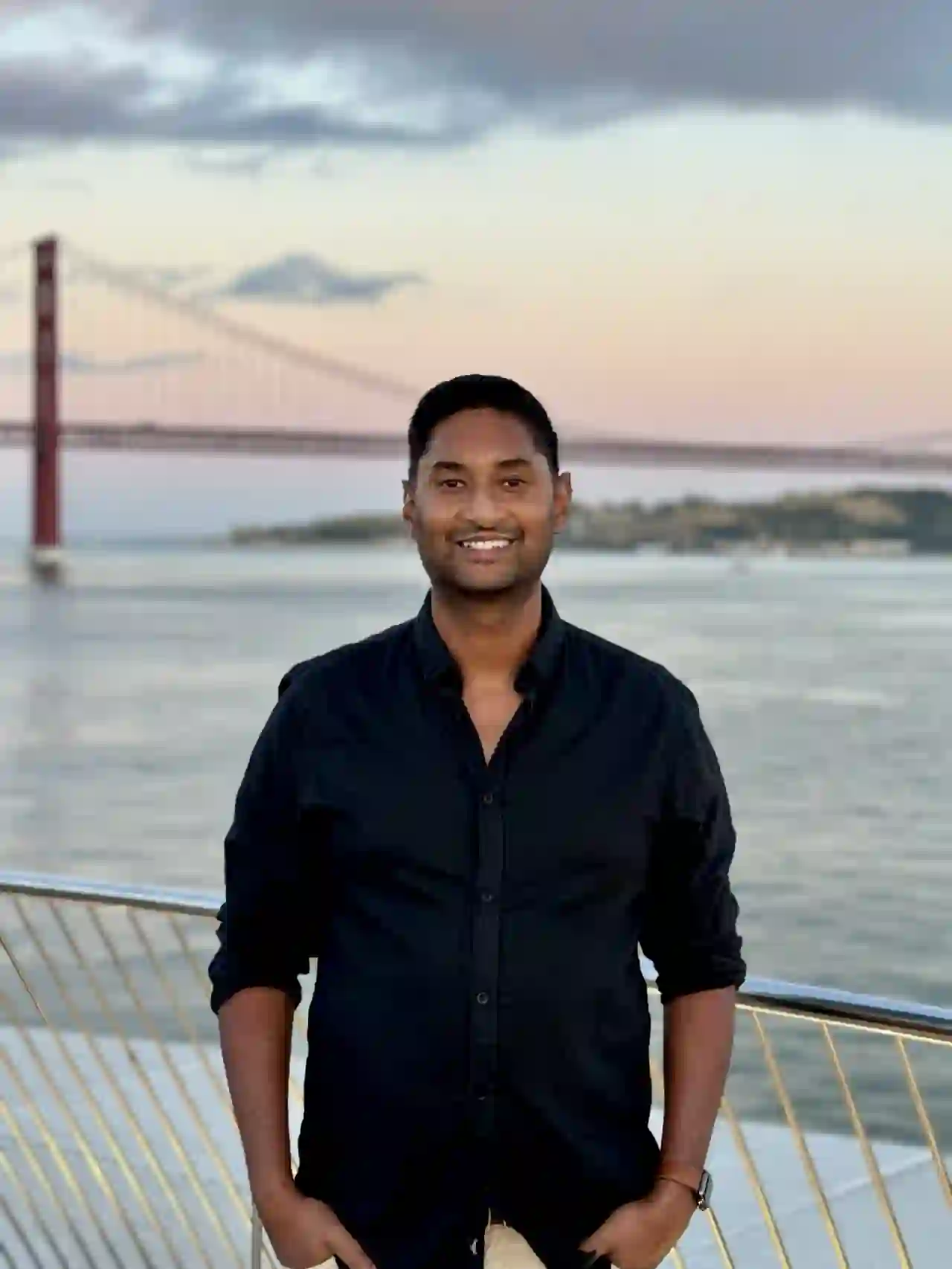 Portrait of Thivanka De Silva smiling outdoors by a river with a suspension bridge in the background